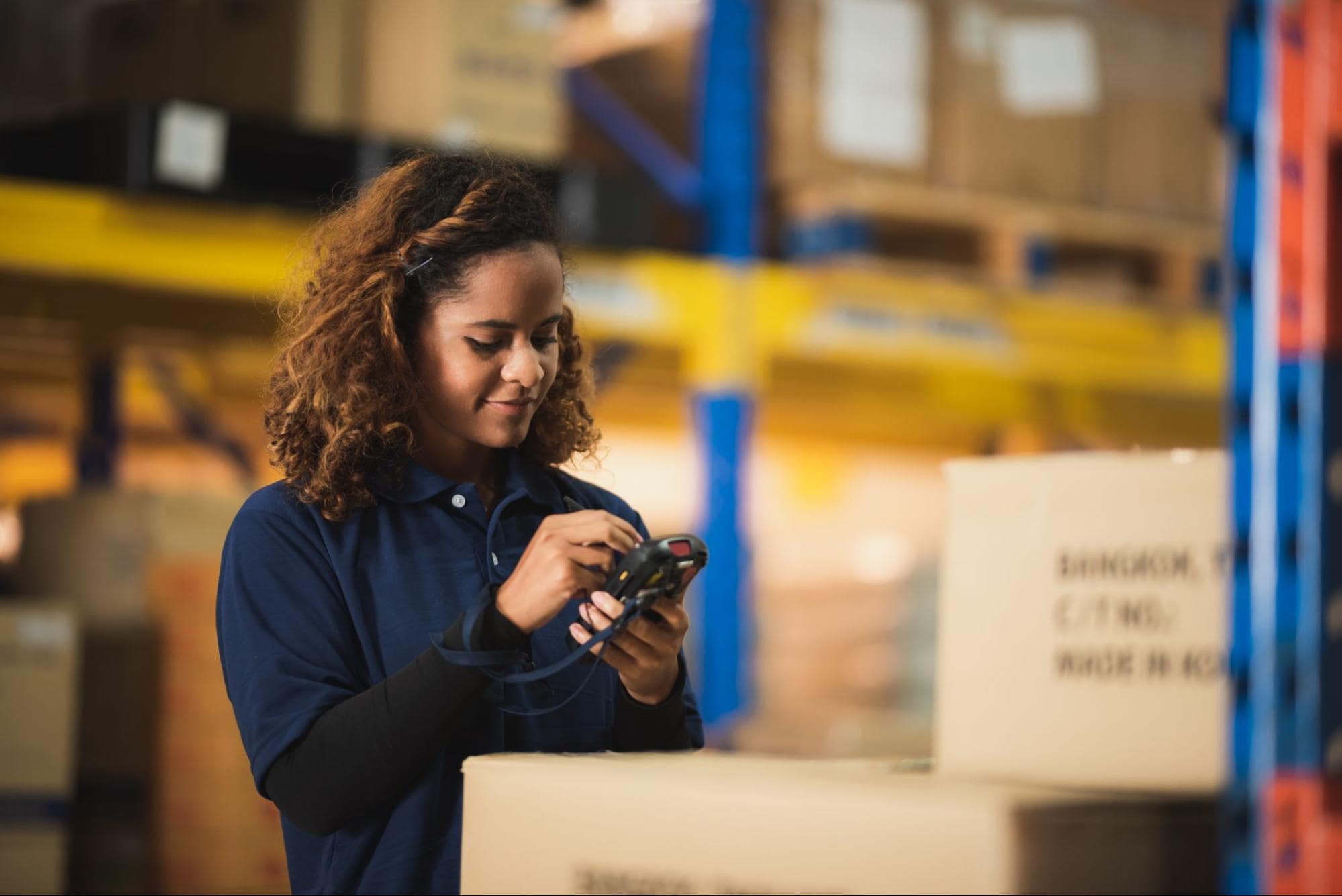 A female warehouse worker operating a handheld picking device standing next to cardboard boxes.