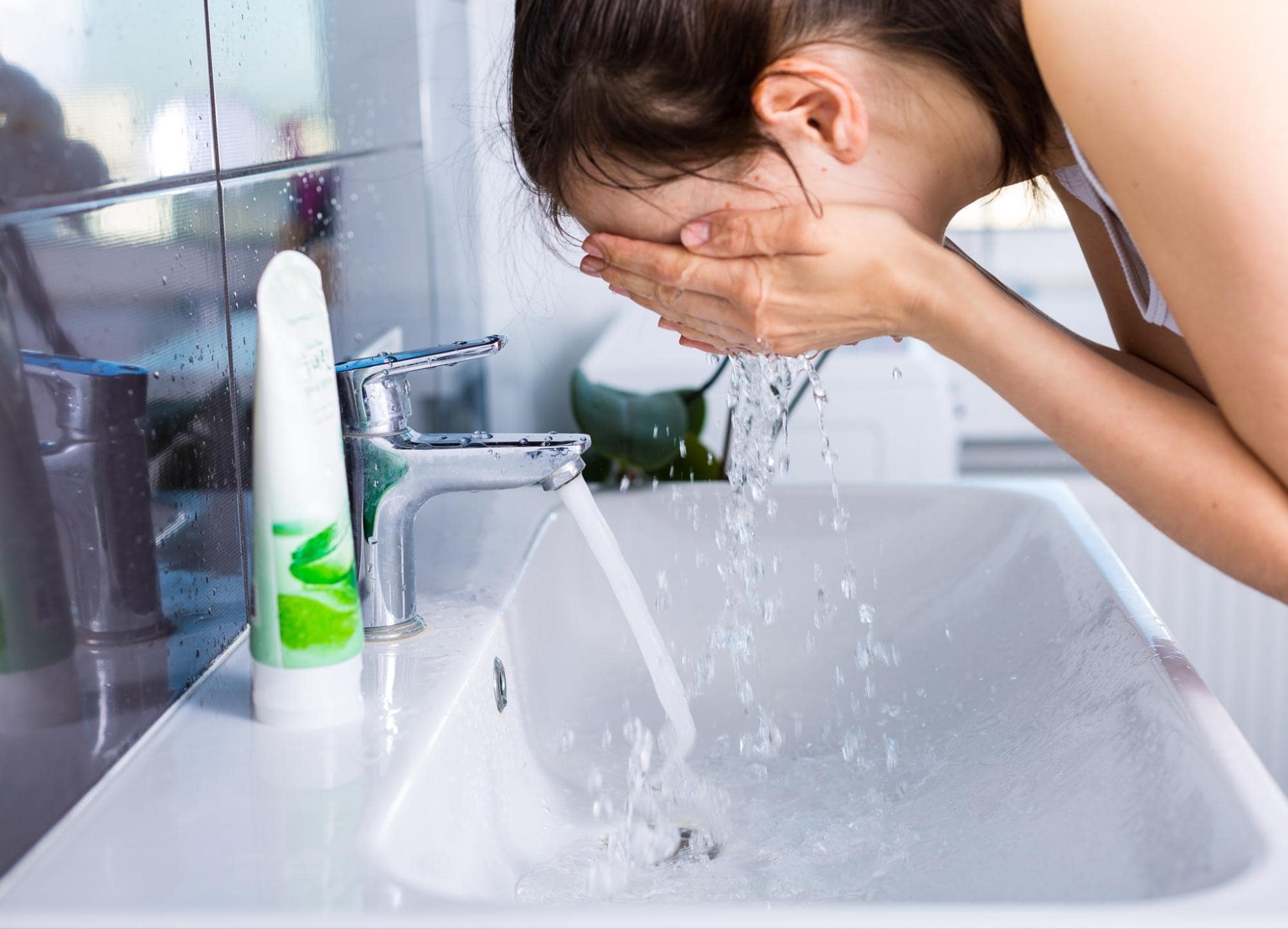 A woman washing her face in the bathroom sink with a face wash tube next to her.