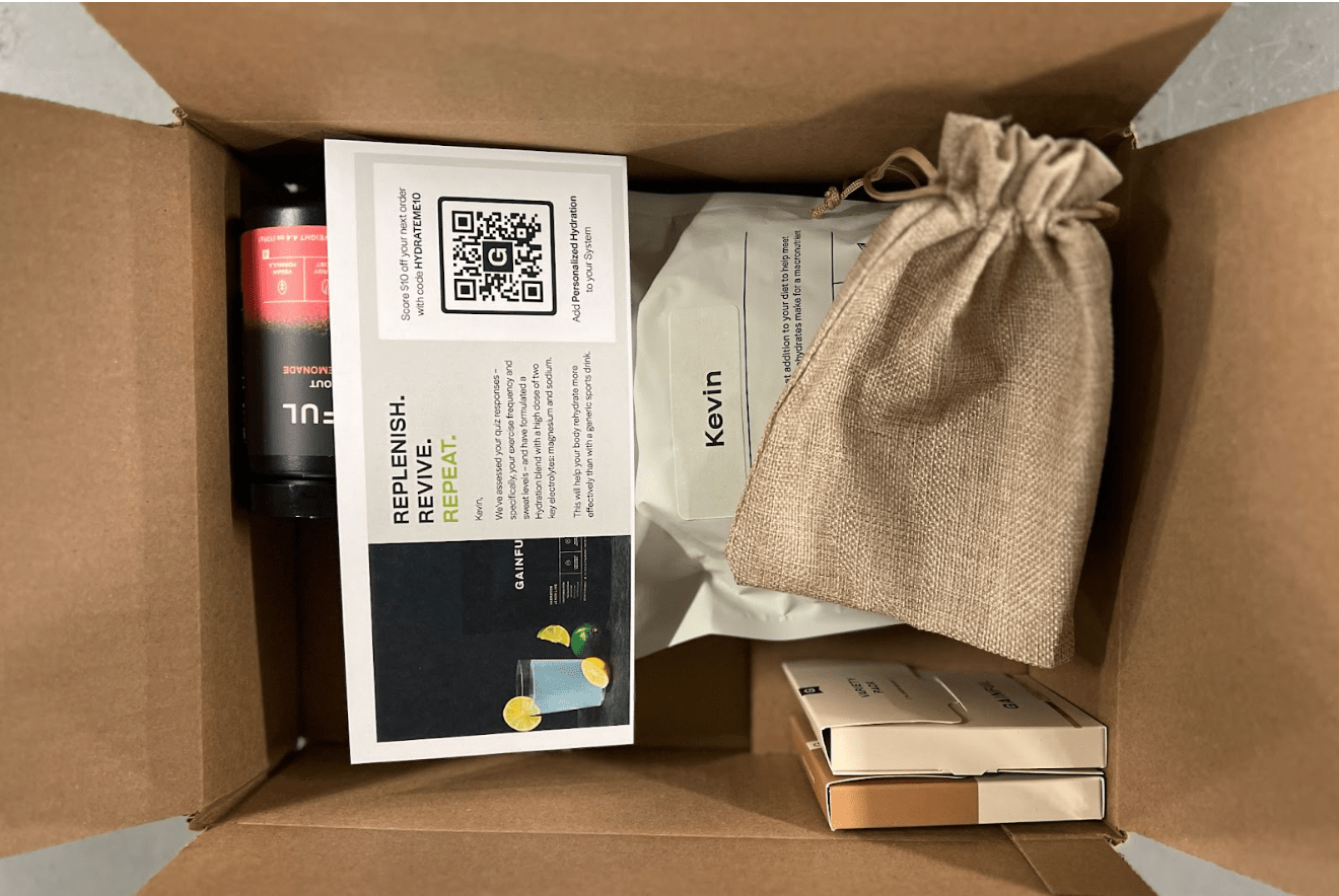 An opened cardboard box with an eCommerce shipment inside. Items include a gift bag and nutrition supplements.
